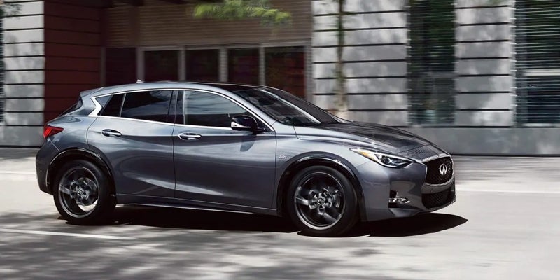 Side view of a dark gray Infiniti crossover driving on a city street with buildings in the background.