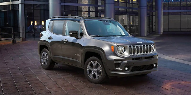 A charcoal gray Jeep Renegade with a black roof parked on a tiled surface in front of a modern building.