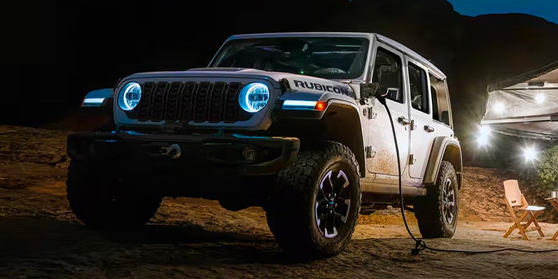 A white Jeep Rubicon 4xe with glowing blue headlights is parked in a dark, rocky area, charging.