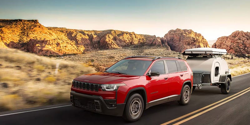 Red SUV towing a teardrop camper on a desert highway with rocky hills.