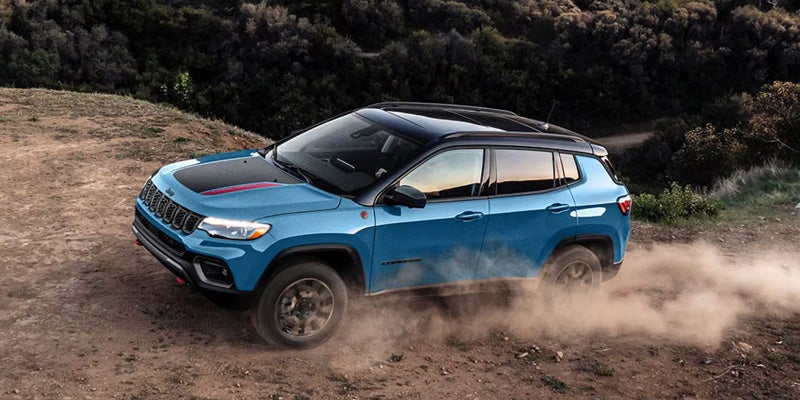 A blue Jeep Compass driving on a dirt road, kicking up dust, with trees in the background.