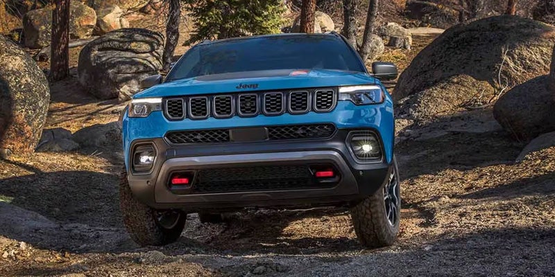 Front view of a blue Jeep Compass Trailhawk on a rocky dirt path surrounded by trees and boulders.