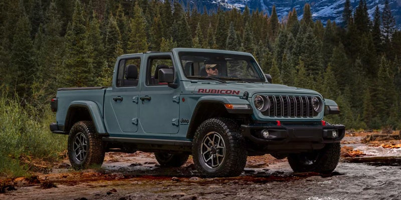 Light bluish-green Jeep Gladiator pickup truck, driven by a man wearing sunglasses, on a dirt path next to a river.