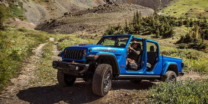 A blue Jeep Gladiator Mojave pickup truck with no doors or roof, driven by a man, on a dirt path in mountains.