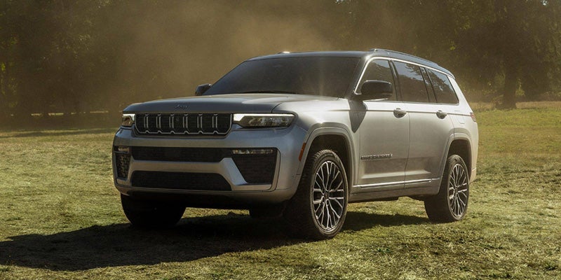 A silver Jeep Grand Cherokee L SUV parked on a grassy field with trees in the hazy background.