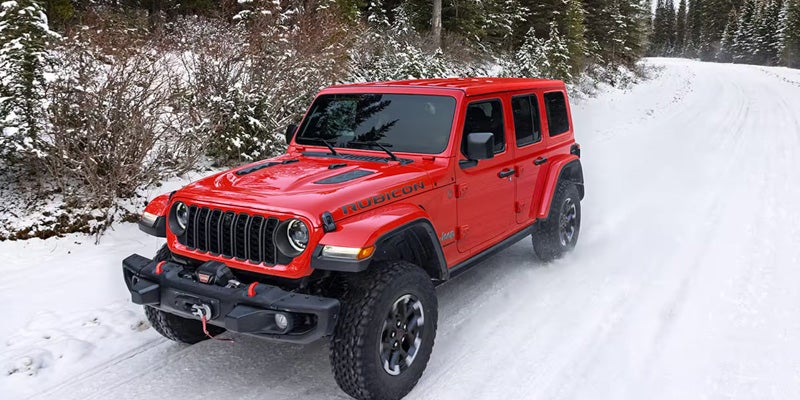 Jeep Wrangler Rubicon driving on a snow-covered road lined with evergreen trees.