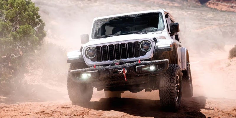 A white Jeep driving off-road on a rocky trail, kicking up dust with its wheels