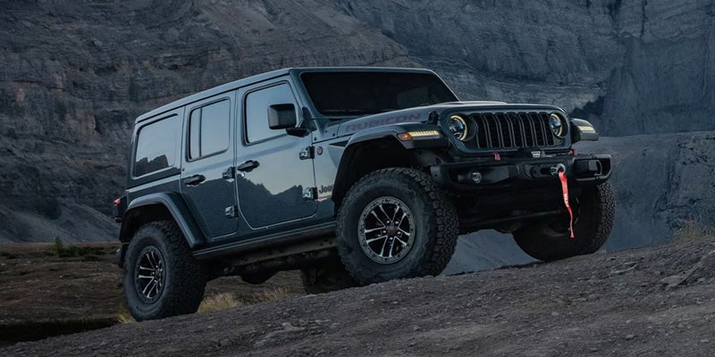 Jeep Wrangler Rubicon with red tow hooks climbing a dirt hill against a rocky, dark background.