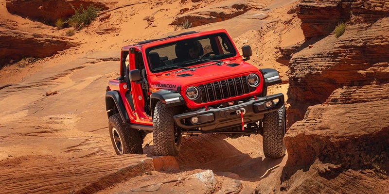 Jeep Wrangler Rubicon off-roading on rocky, orange-brown terrain with its front left wheel lifted up.
