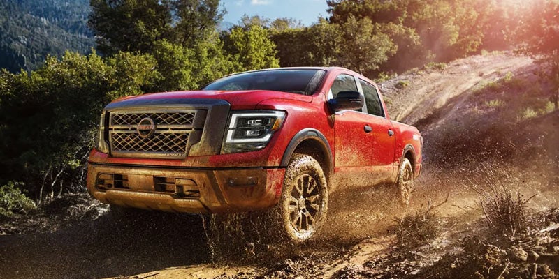 A red pickup truck splashes through mud on an off-road trail surrounded by green trees under a bright sky.