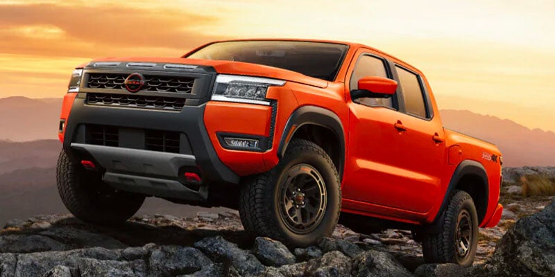 Orange Nissan Frontier Pro-4X pickup truck on rocky terrain with mountains and a golden sky in the background.