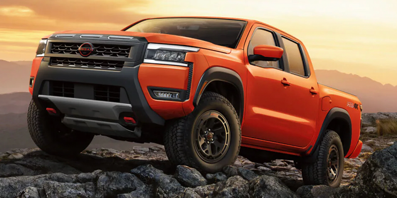 An orange Nissan Frontier pickup truck climbs over rocks with mountains and an orange sky in the background.