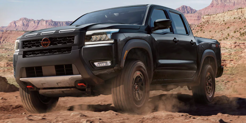 A black Nissan Frontier pickup truck drives across a dirt road with red rock formations in the background.