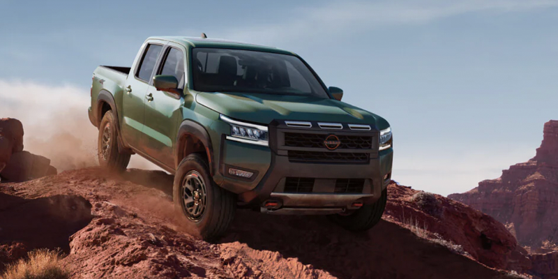 A green Nissan Frontier pickup truck drives uphill on a dirt road in a desert landscape with red rock formations.