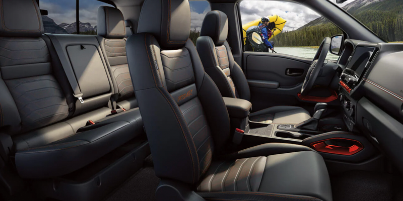 View from inside a car: black leather interior with orange stitching, mountains and river with kayaker visible outside.
