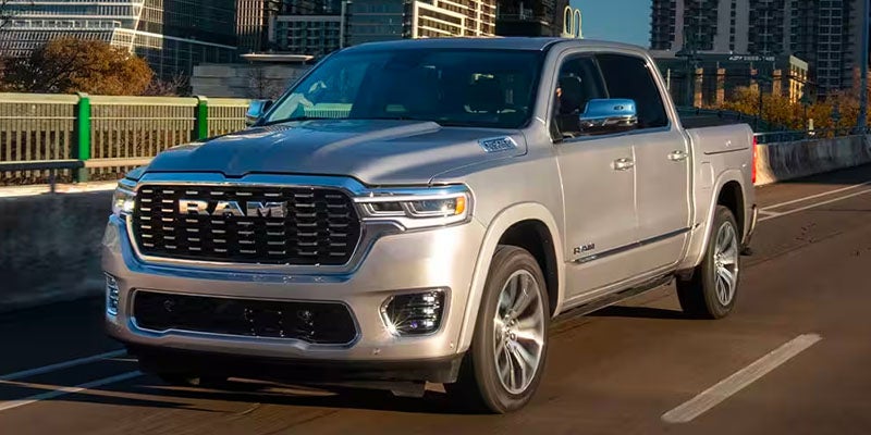 A silver Ram pickup truck with chrome accents drives on a highway overpass, partially obscuring city buildings.