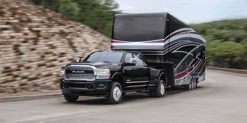 Black RAM 3500 pickup truck towing a large black fifth-wheel trailer on a winding road with a stone wall.