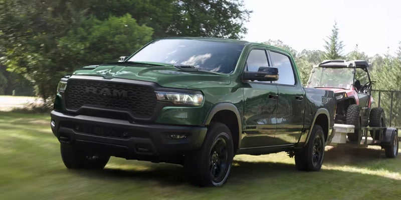 Ram pickup truck drives away on a grassy lawn, pulling a red UTV on a trailer.