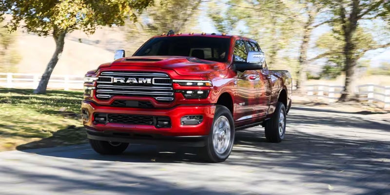 A shiny red Ram pickup truck with chrome accents drives on a paved road, trees and white fences in the background.