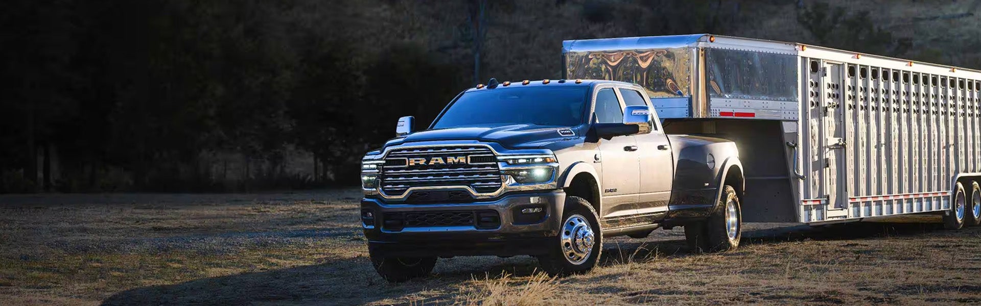A black Ram dually truck pulling a large, silver livestock trailer on a dirt road with dark trees in the background.