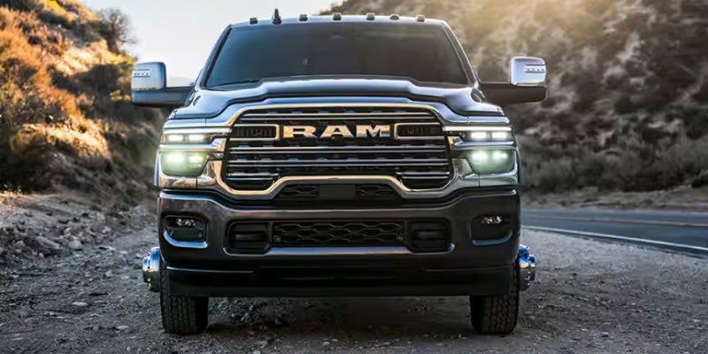 Front view of a dark gray Ram pickup truck with headlights on, parked on a dirt road next to a paved road.