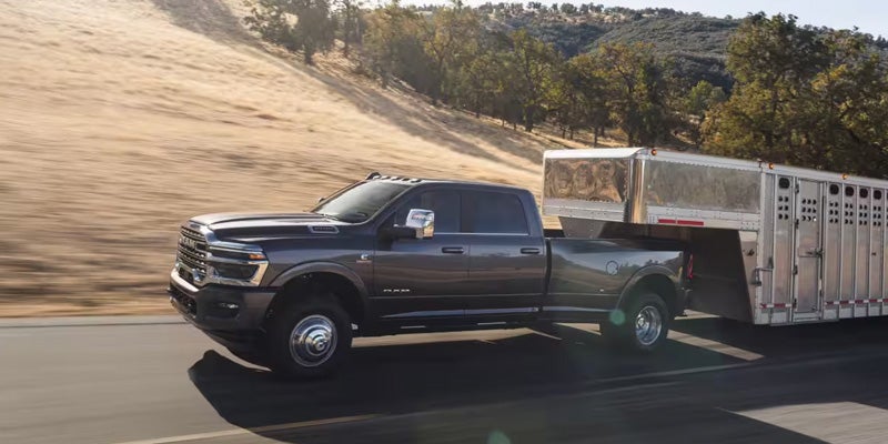 Ram 3500 truck towing a large silver livestock trailer down a highway with rolling golden hills.