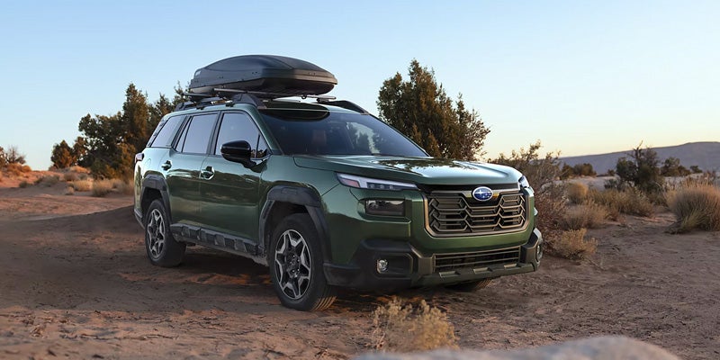 A dark green Subaru SUV with a roof cargo box is parked on a dirt path in a desert landscape