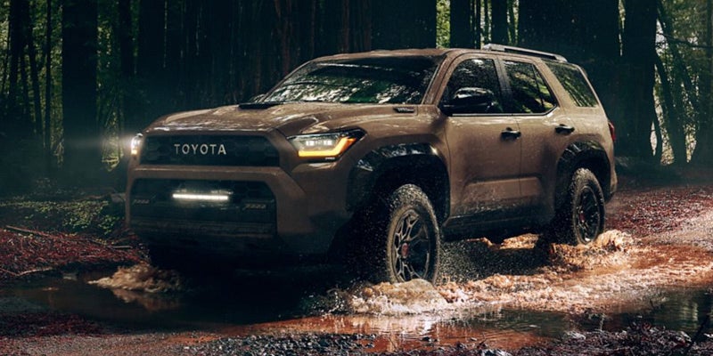 A brown Toyota Sequoia SUV with headlights on splashing through mud and water in a dark forest.