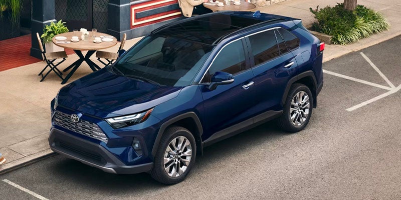 A dark blue Toyota RAV4 SUV parked on asphalt in front of a cafe with outdoor seating.