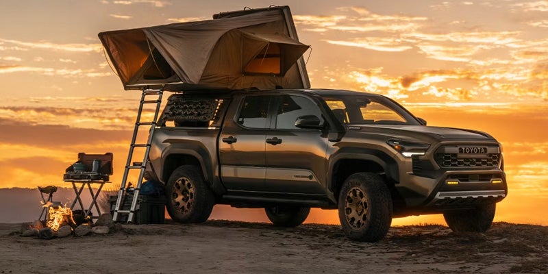 Dark grey Toyota truck with a rooftop tent and ladder, campsite setup with campfire, at sunset.