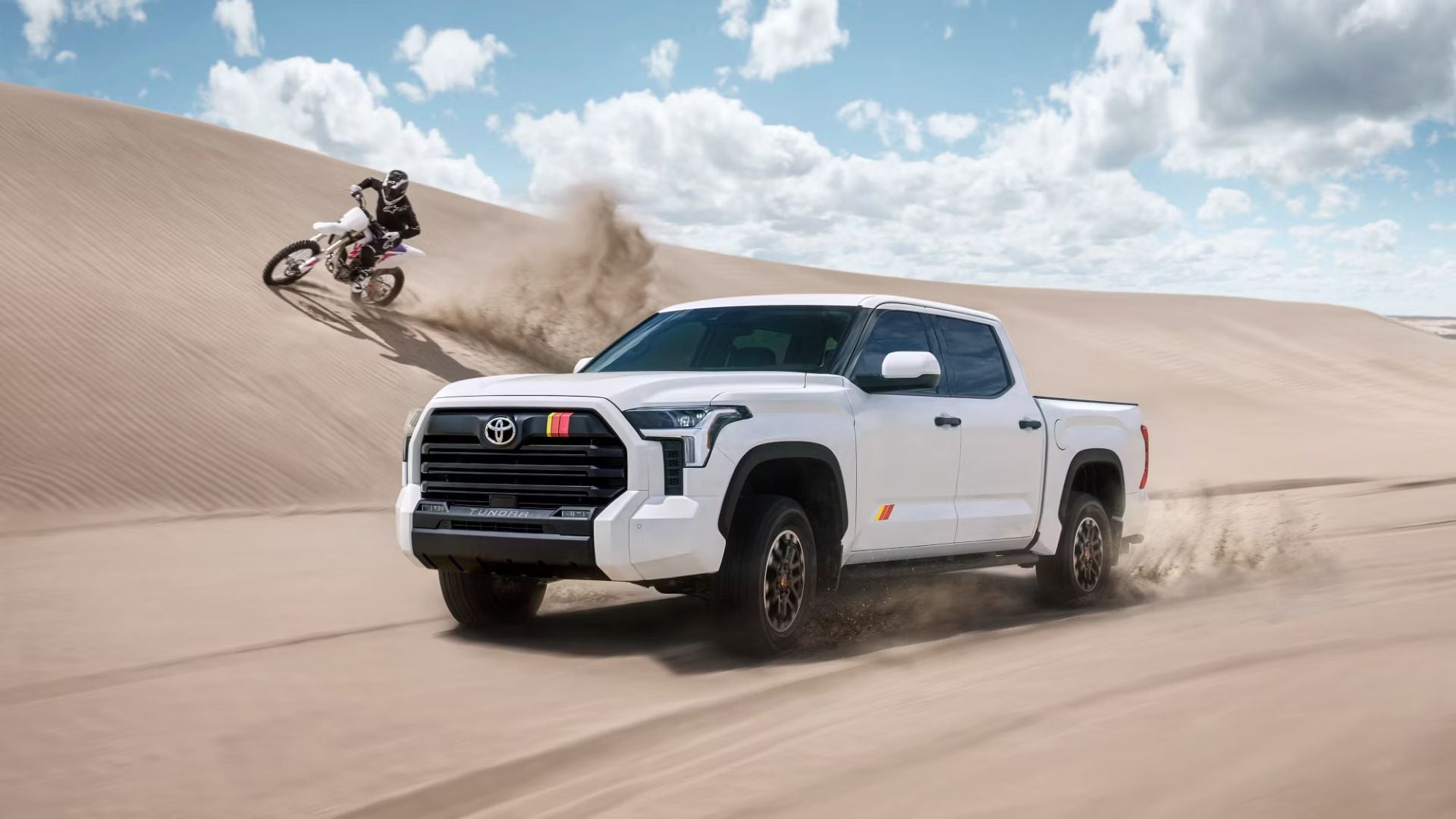 White Toyota Tundra pickup truck drives on a sandy dune, a motocross rider in the background.