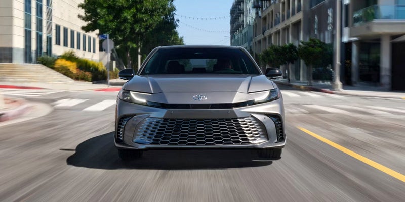 Front view of a gray car with a large, low grille, driving on a city street with blurred buildings.