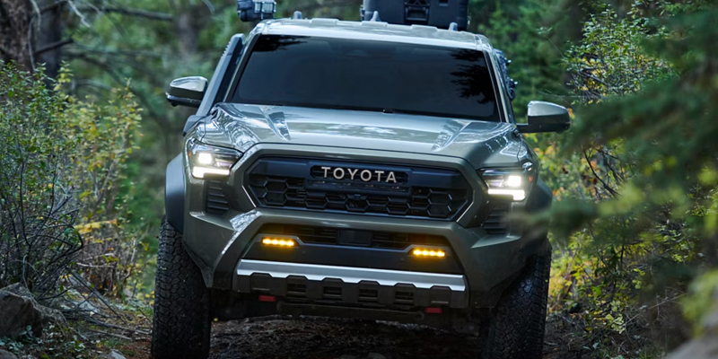 A front view of a dark olive green Toyota truck with its headlights on, driving on a dirt path in a forest.