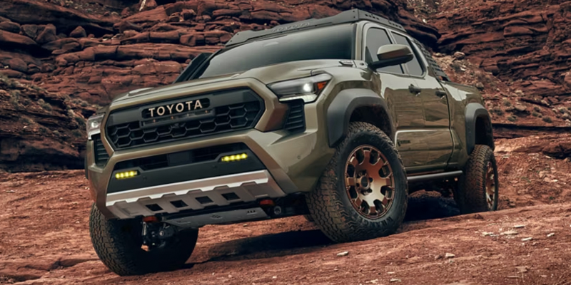 A bright red GMC pickup truck parked on a dirt trail in a sun-dappled forest of tall evergreen treeA green Toyota pickup truck with bronze wheels and extra lights driving on a rocky dirt road.
