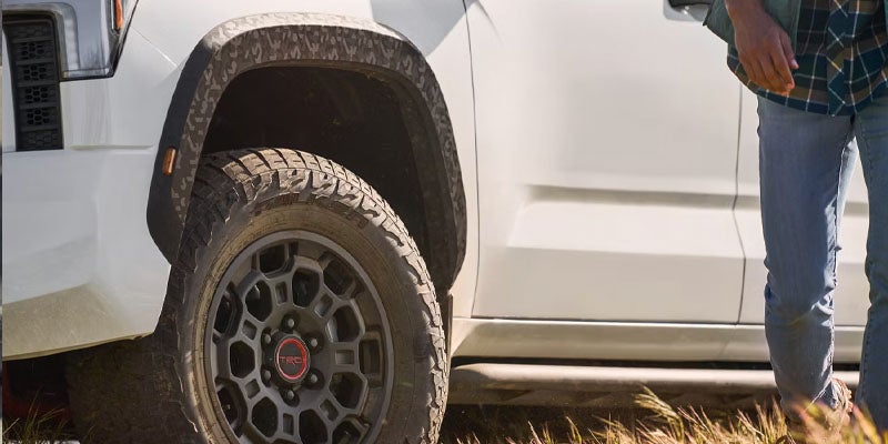 Close-up of a white truck's front wheel with a TRD red and black hub cap and a person's legs nearby.