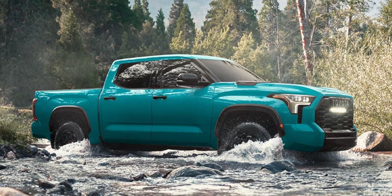 A turquoise Toyota Tundra pickup truck drives through a shallow rocky river in a forest.