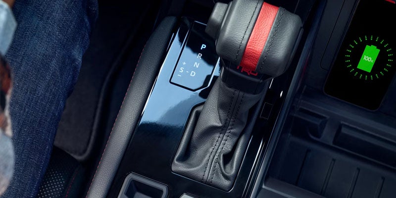 A car's interior features a black and red TRD shift knob, a gear selector, and a 100% charged phone.