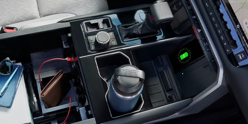 A car's interior with an open center console revealing storage for a wallet, sunglasses, and a phone, plus a water bottle in a cup holder and a wireless charger.