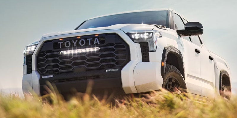 Frontal view of a white Toyota Tundra with an illuminated light bar on the grille, driving through tall grass.