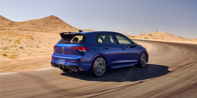 A blue Volkswagen Golf R drives away on a desert road with mountains in the background.