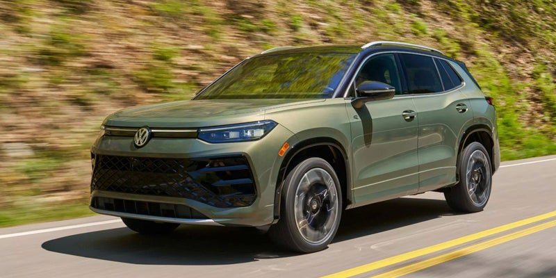 Volkswagen Taos SUV with a black roof and silver roof rails drives on a highway next to a green hill.