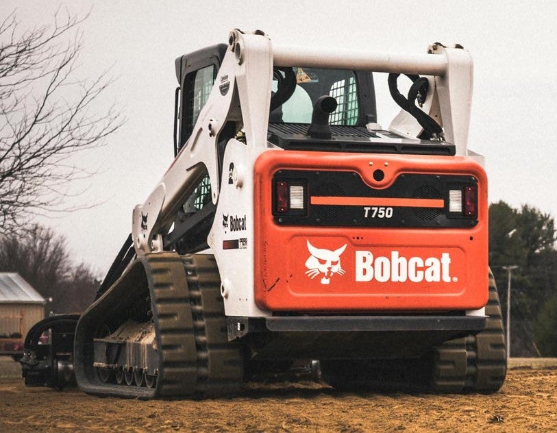 Rear view of a Bobcat T750 compact track loader parked on a dirt field with trees in the background.
